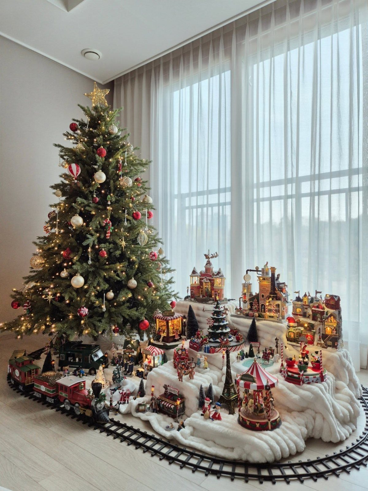 90s Christmas Tree Decor: 8 Ideas for Retro Holiday Charm 3 A miniature Christmas village with illuminated houses and a carousel sits beside a decorated tree.