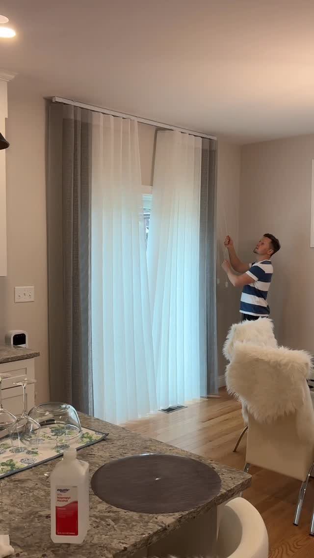 A man adjusts long, sheer white and grey curtains hanging from a rod over a large window.