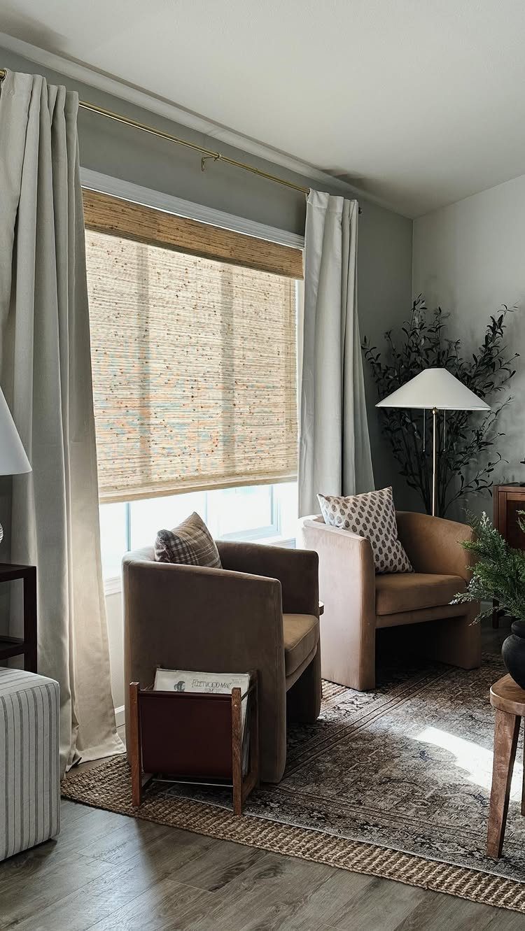 Woven wood shades hanging in a window framed by light grey drapery curtains in a living room.