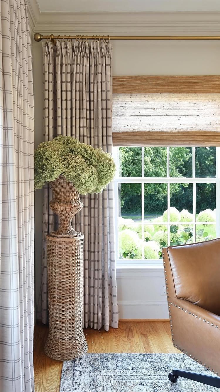 Bamboo shade and patterned curtains hanging over a window next to a wicker pedestal with flowers.