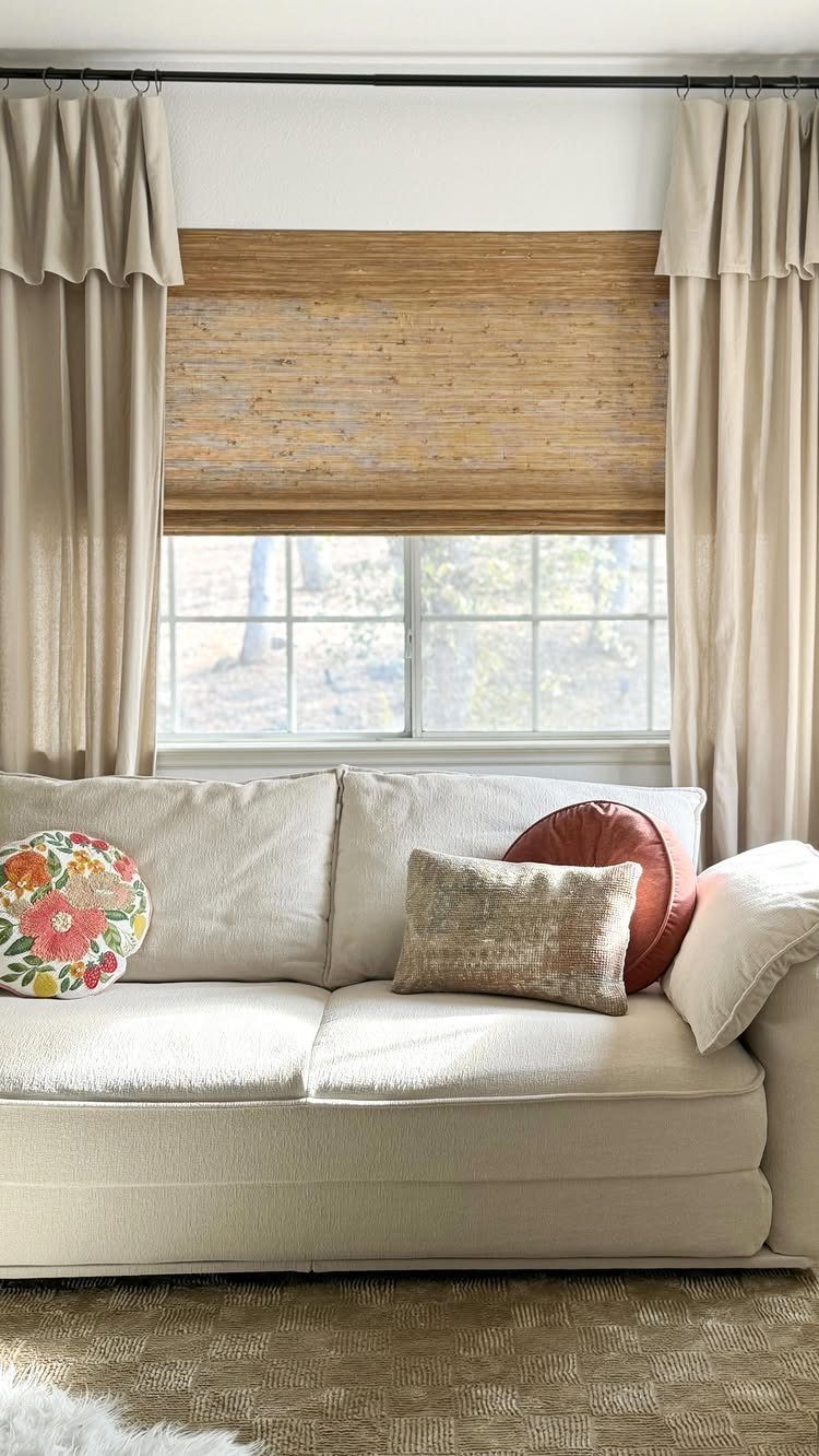 Woven bamboo shade hanging over a window flanked by long beige curtains and a cream sofa.