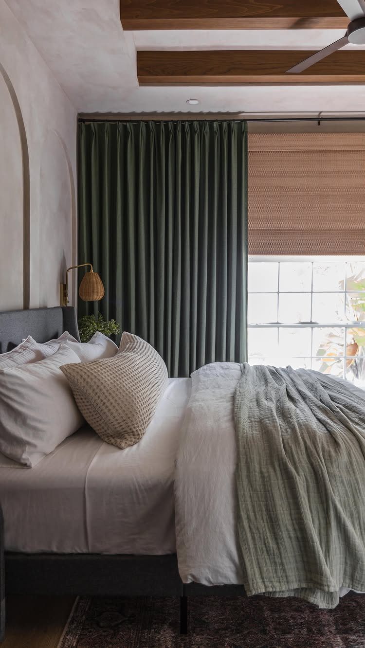 10 Bedroom Window Decor Ideas for Every Wall Height 5 A bed with white linens sits beside a window with green curtains and a Roman blind.