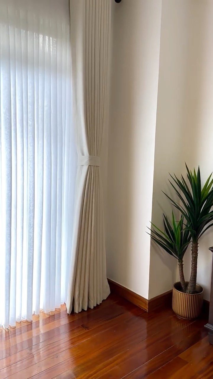 10 Bedroom Window Decor Ideas for Every Wall Height 4 White sheer curtains hanging by a window next to a potted green plant on wooden flooring.