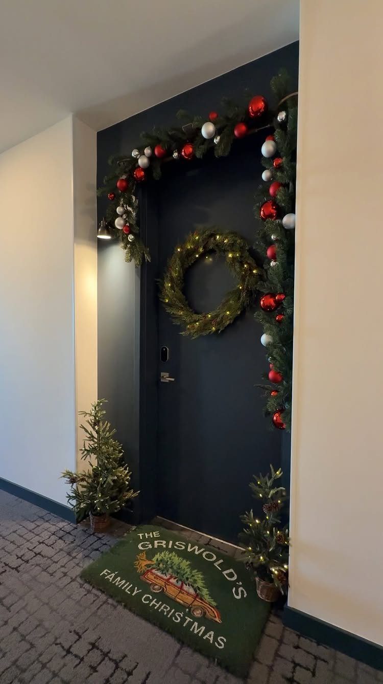 25 Christmas Decor Apartment Ideas That Maximize Space 23 A dark apartment door decorated with Christmas garlands, a wreath, small trees, and a themed doormat.