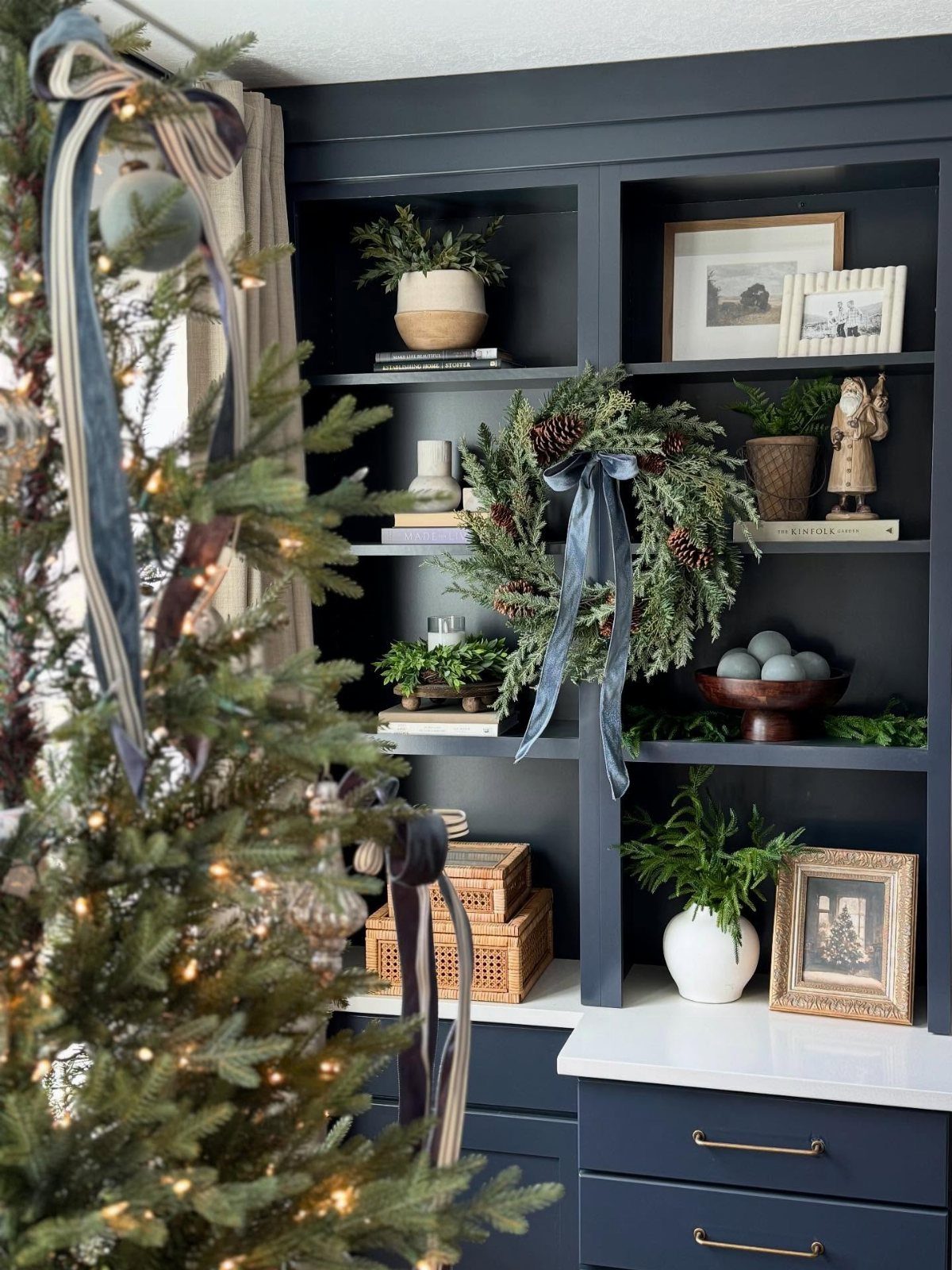 25 Christmas Decor Apartment Ideas That Maximize Space 19 A decorated Christmas tree with blue ribbons stands in front of a dark blue bookshelf.
