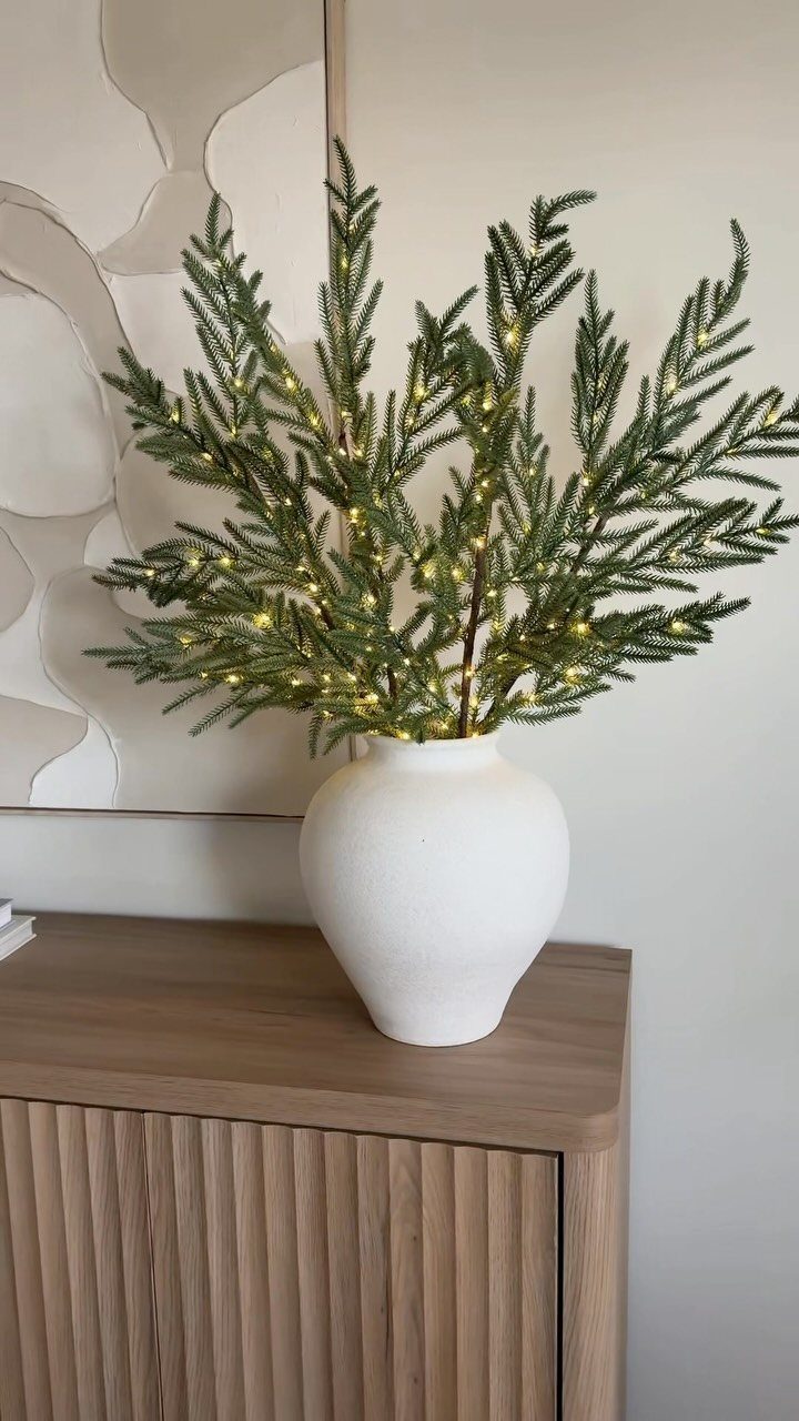 21 Christmas Decor Inspiration Ideas for Your Whole Home 2026 11 Evergreen branches with warm twinkle lights arranged in a white vase on a wooden cabinet.