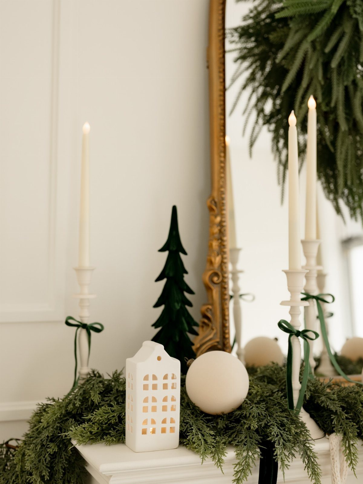 21 Christmas Decor Inspiration Ideas for Your Whole Home 2026 12 White taper candles and green garland decorate a mantel with neutral ornaments and a small ceramic house.