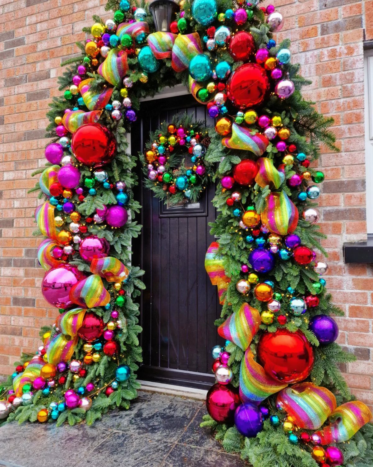 21 Christmas Decor Inspiration Ideas for Your Whole Home 2026 14 A dark door framed by a lush evergreen garland covered in large, colorful jewel-toned Christmas ornaments.