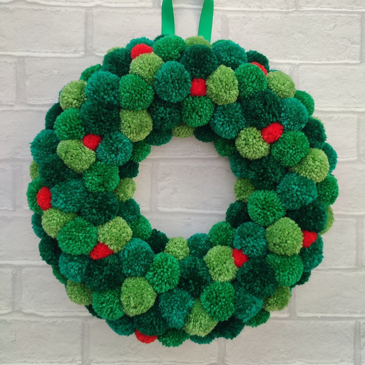21 Christmas Decor Inspiration Ideas for Your Whole Home 2026 3 A handmade wreath composed of green and red yarn pom-poms hanging on a white brick wall.