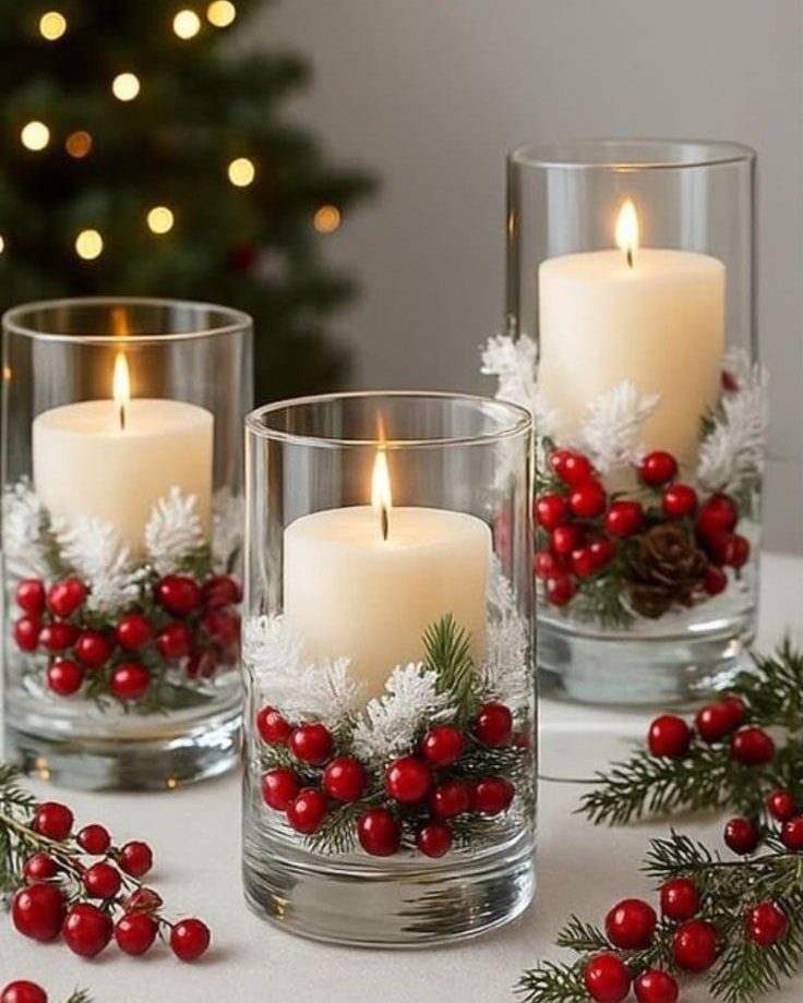 Three lit white pillar candles in glass holders decorated with red berries and frosted pinecones.