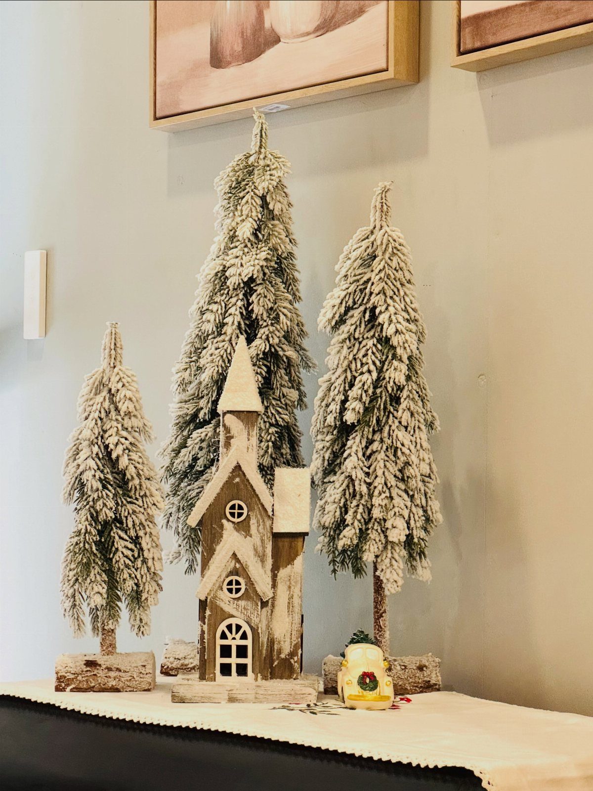 Frosted evergreen trees and a small wooden house figurine arranged on a mantelpiece.