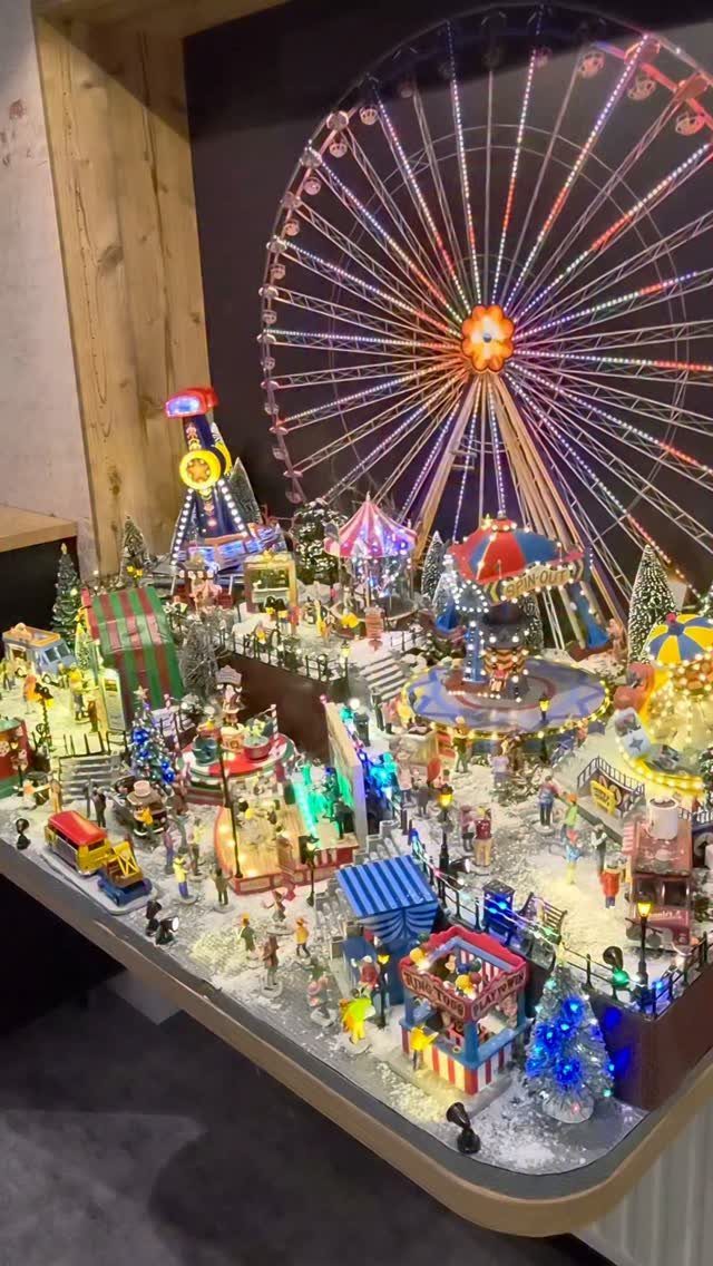 19 Christmas Village Ideas for Displaying a Miniature Scene 19 A miniature carnival-themed Christmas village display with colorful lights and a large Ferris wheel backdrop.