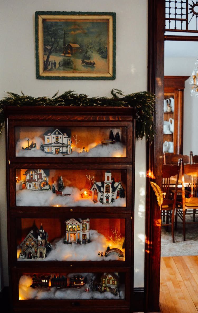 19 Christmas Village Ideas for Displaying a Miniature Scene 5 A wooden curio cabinet displaying a miniature snowy Christmas village with lighted houses on multiple shelves.