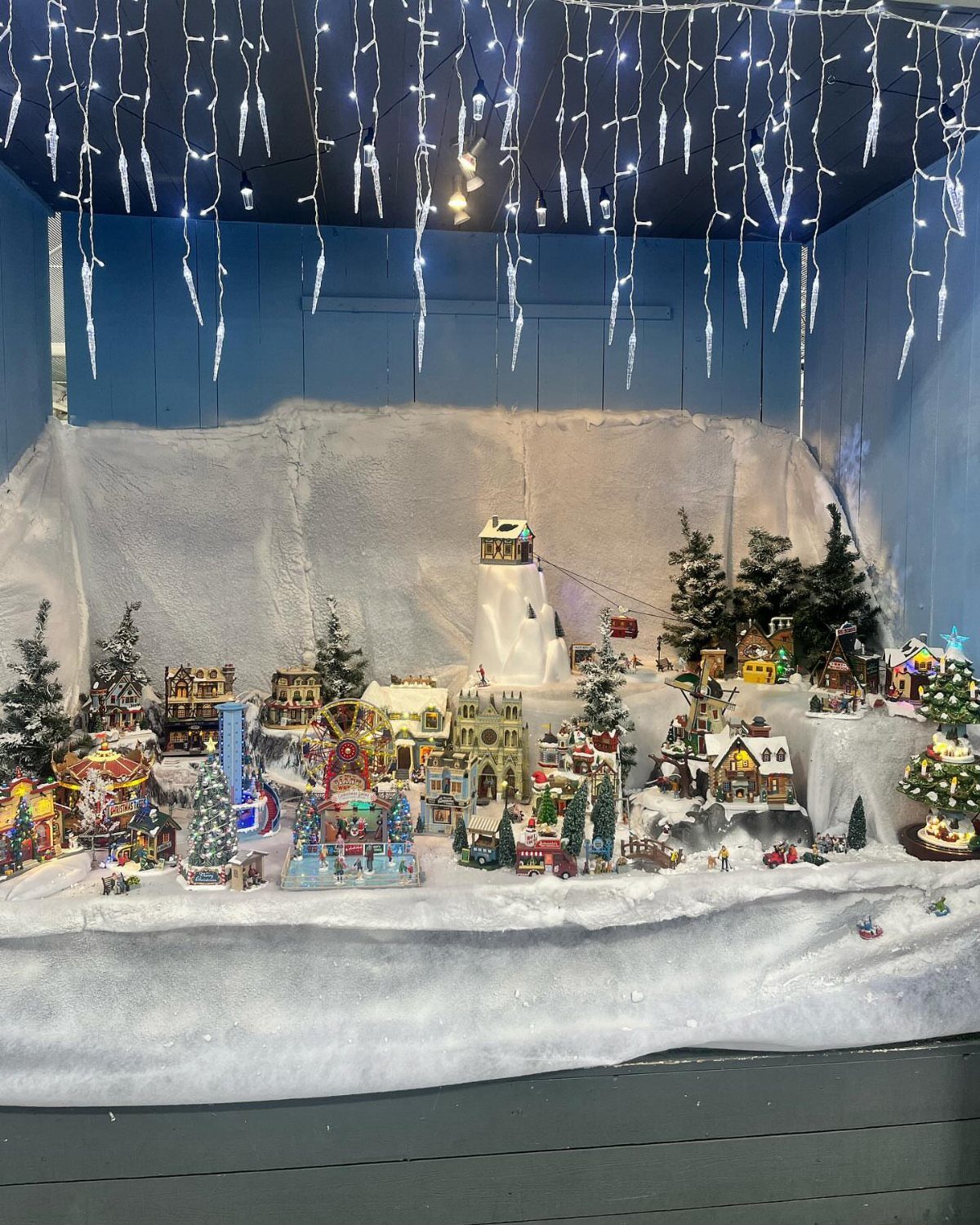 19 Christmas Village Ideas for Displaying a Miniature Scene 9 Miniature Christmas village houses and trees set against a white fabric snowy mountain backdrop.