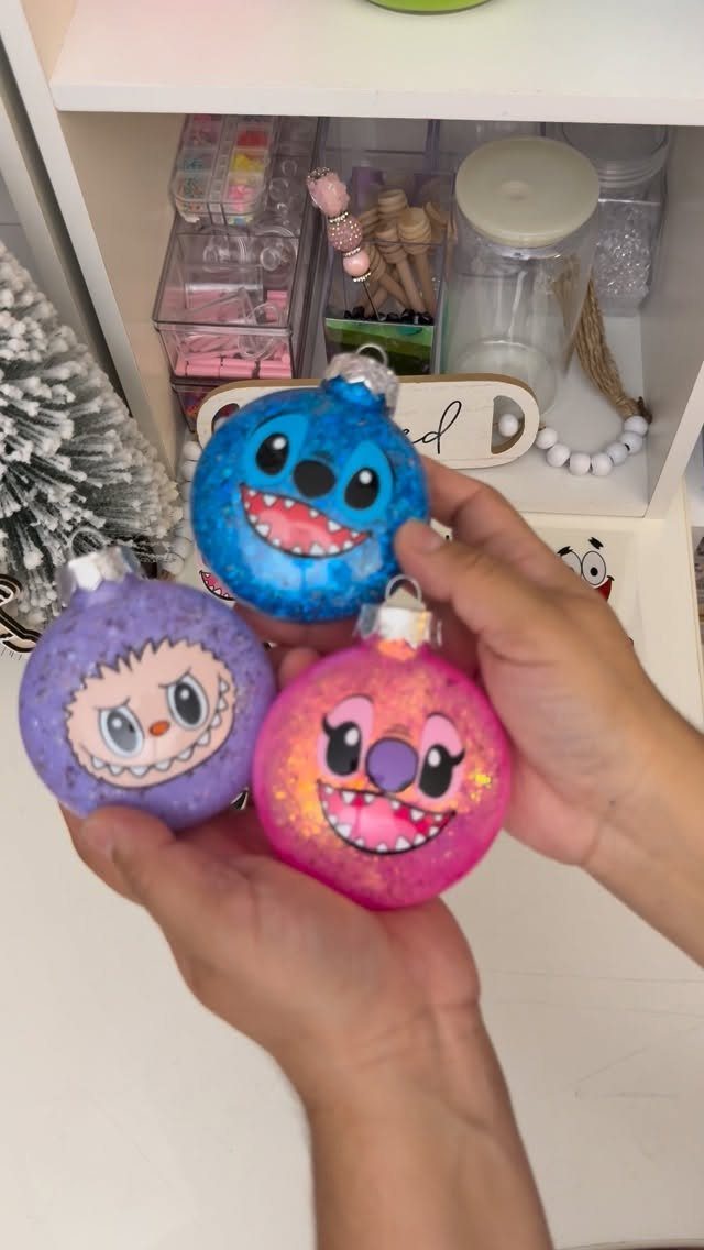 Hands holding three colorful, glittery character-themed Christmas ball ornaments.