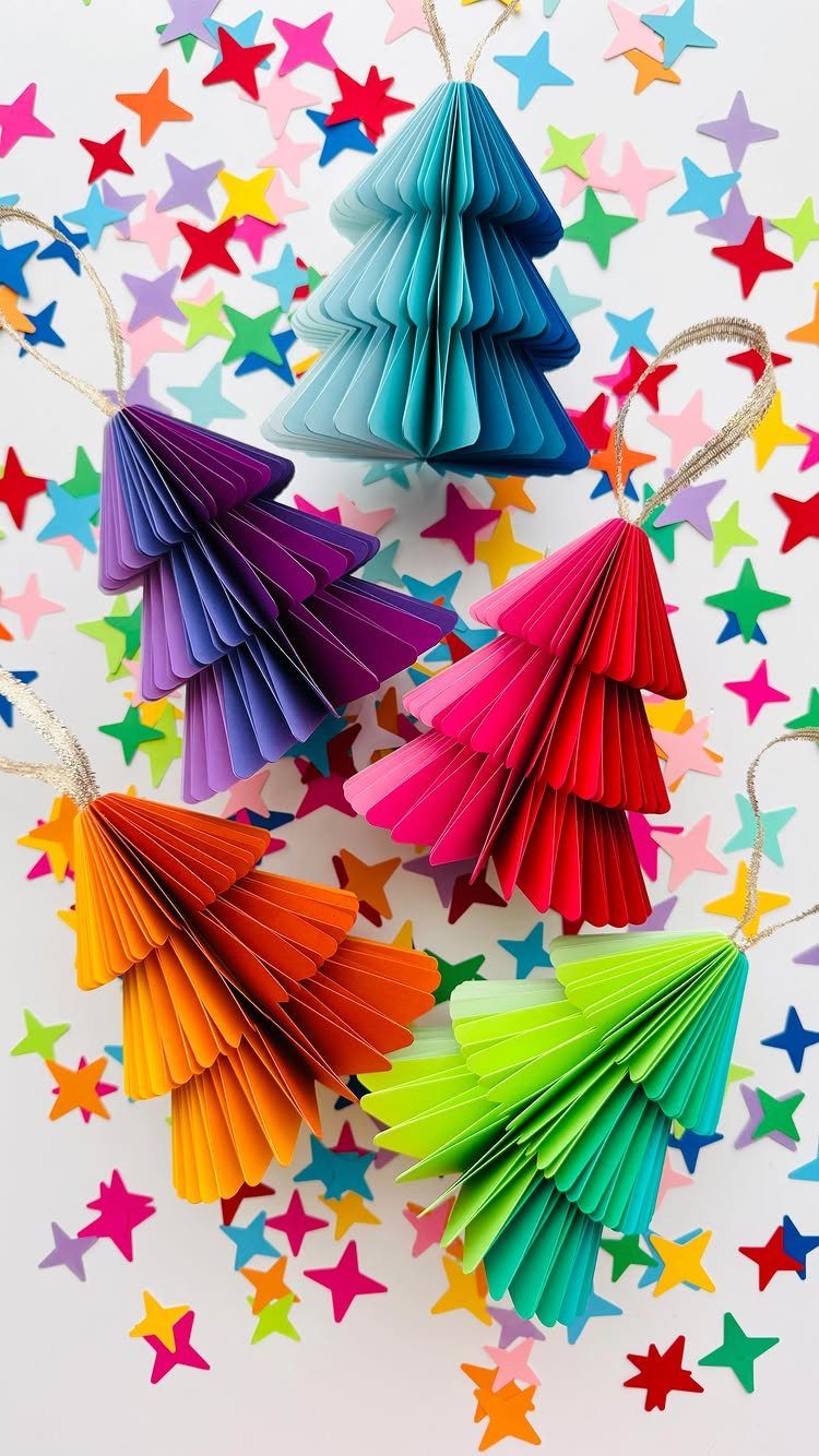 Five colorful accordion-folded paper tree ornaments scattered among small multi-colored star confetti.