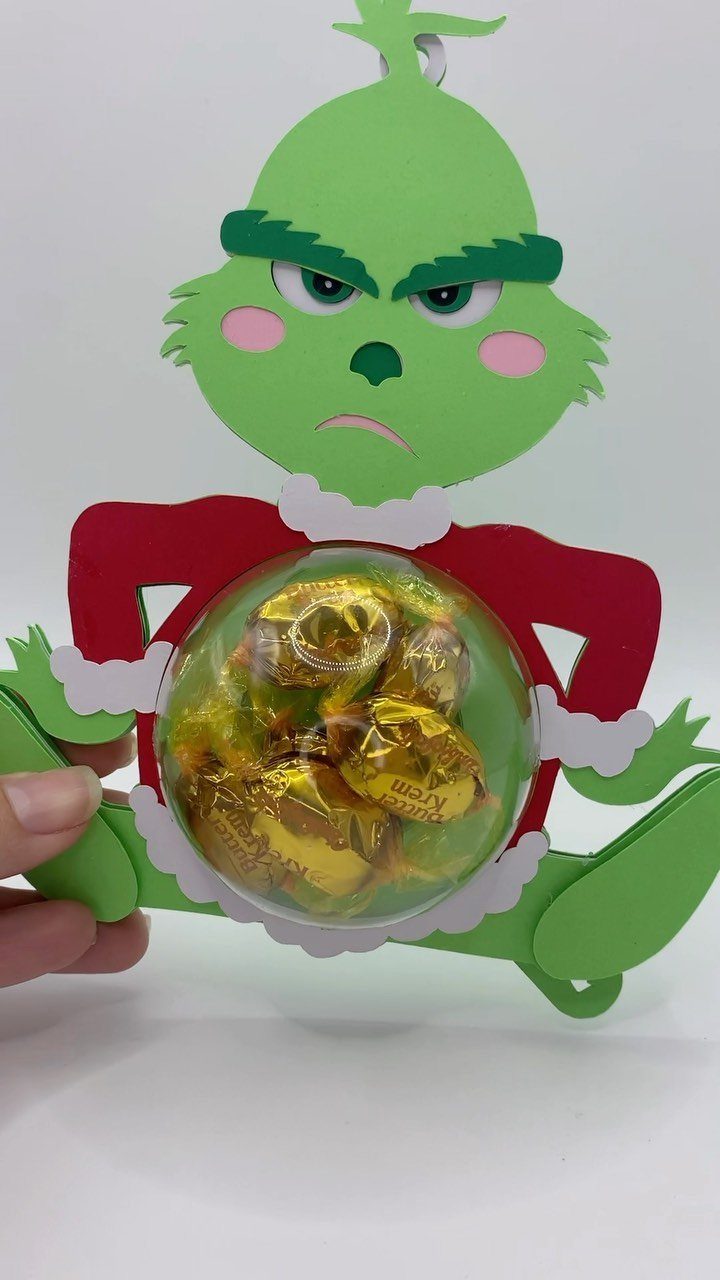 A green Grinch cardstock ornament holding a clear sphere filled with gold wrapped candies.