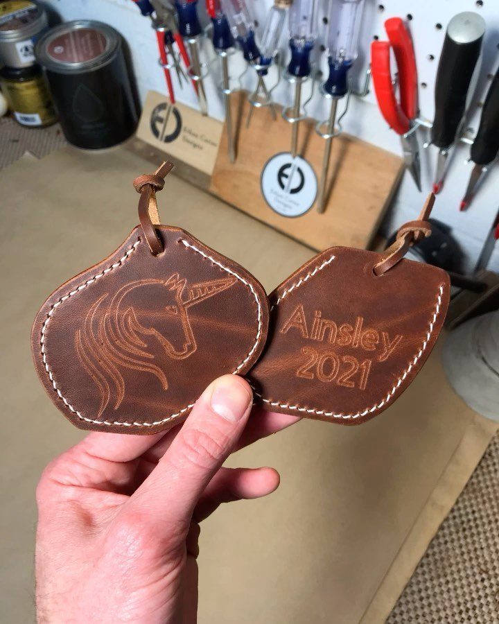 A hand holding two brown leather ornaments with etched unicorn and name designs.