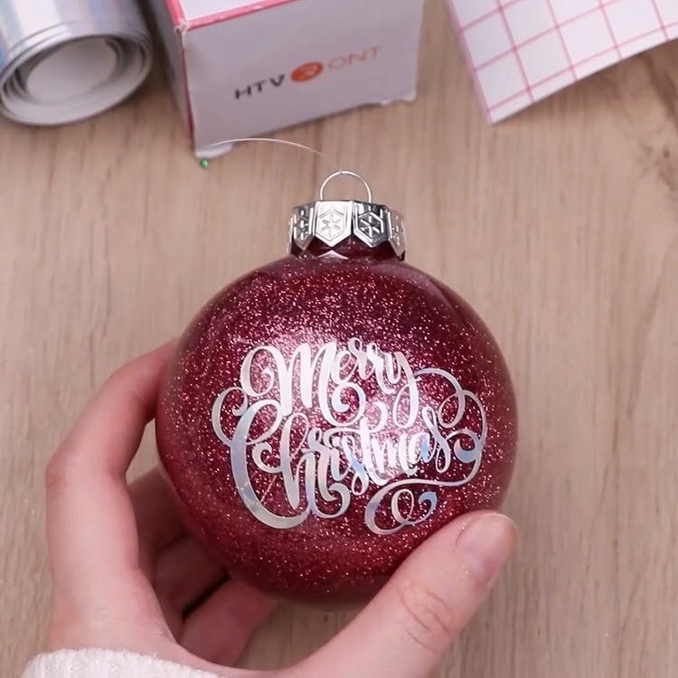 A hand holding a red glittery Christmas ornament with white Merry Christmas script lettering.