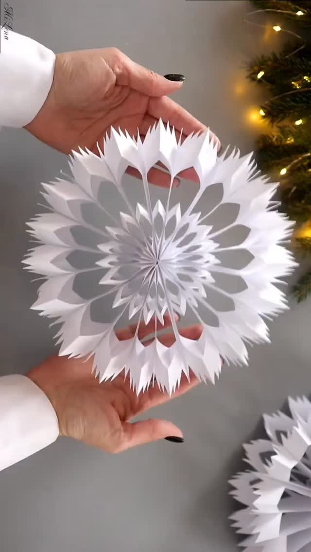 Hands holding a large, intricate white 3D paper snowflake ornament against a blurred background.