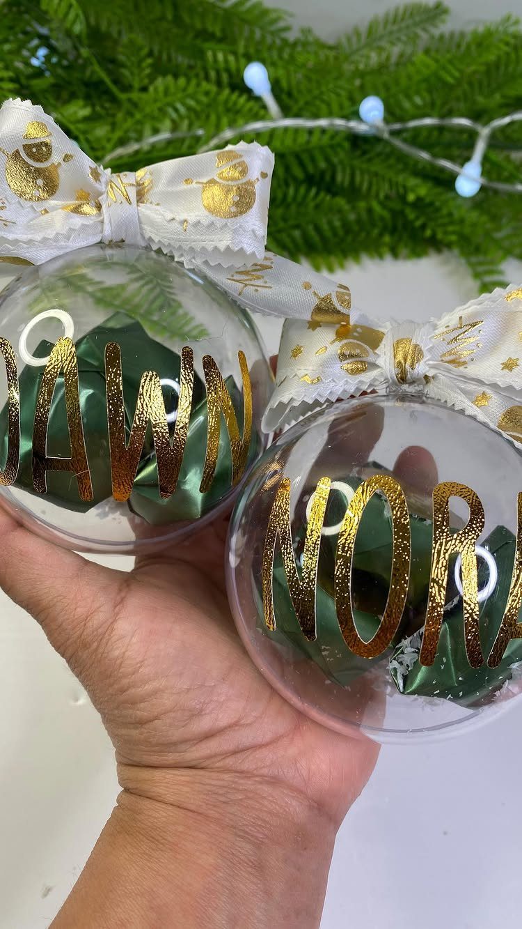 A hand holding two clear plastic Christmas ornaments with gold glitter names and white bows.