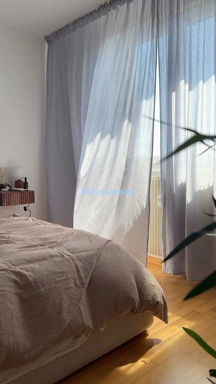 Light blue sheer curtains hanging over a window in a sunlit bedroom with a bed.