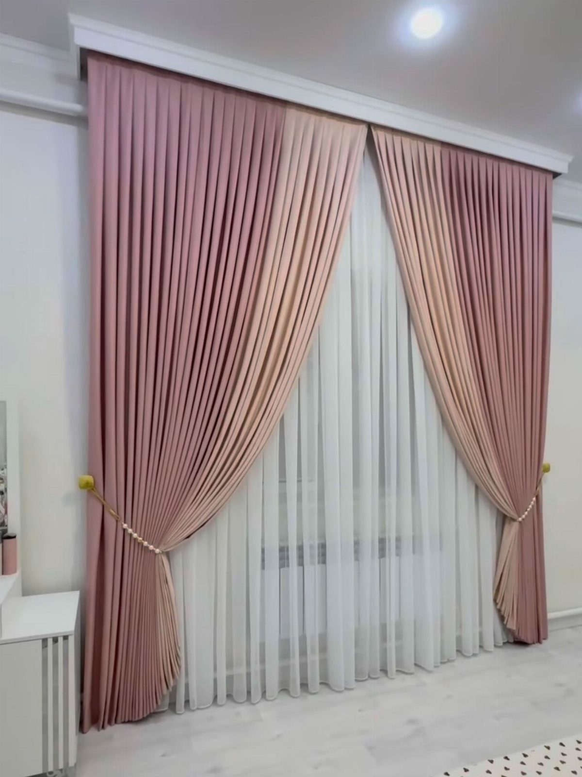 Blush pink pleated curtains draped with gold tie-backs over sheer white window curtains.