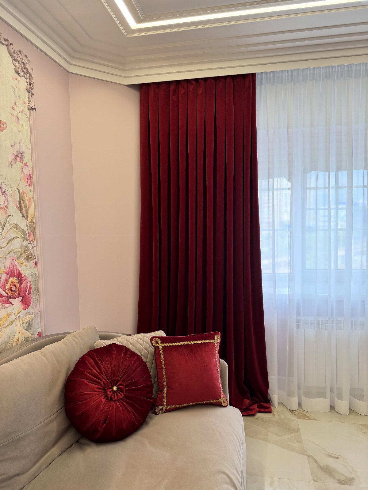 Red velvet curtains hanging next to sheer white curtains in a room with a beige sofa.