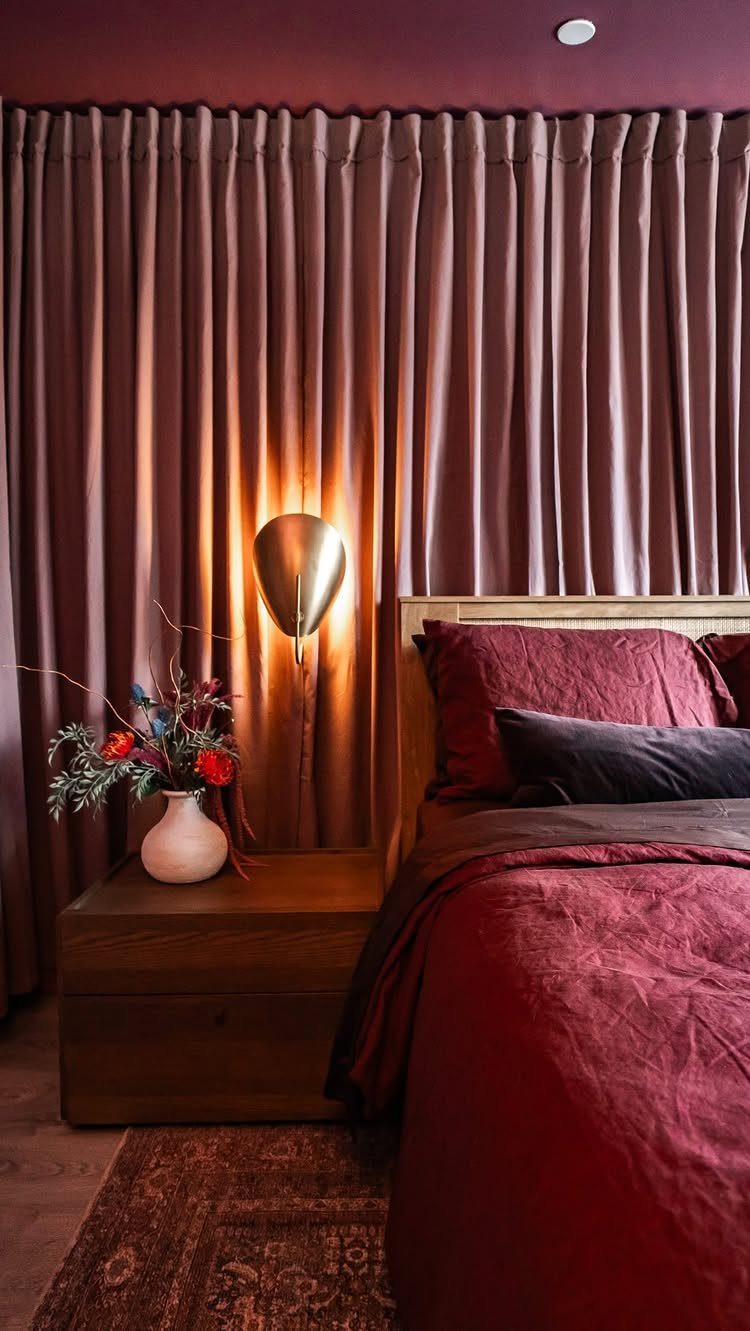 Mauve curtains hang in deep folds behind a bed and a wooden nightstand with flowers.