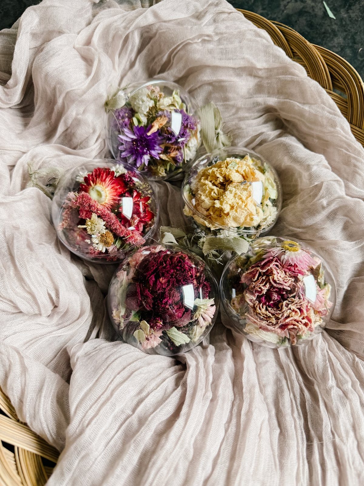 7 Ideas for Decorating Clear Ornaments with Glitter, Crayon & More 4 Five clear glass ornaments filled with colorful dried flowers resting on draped beige fabric.