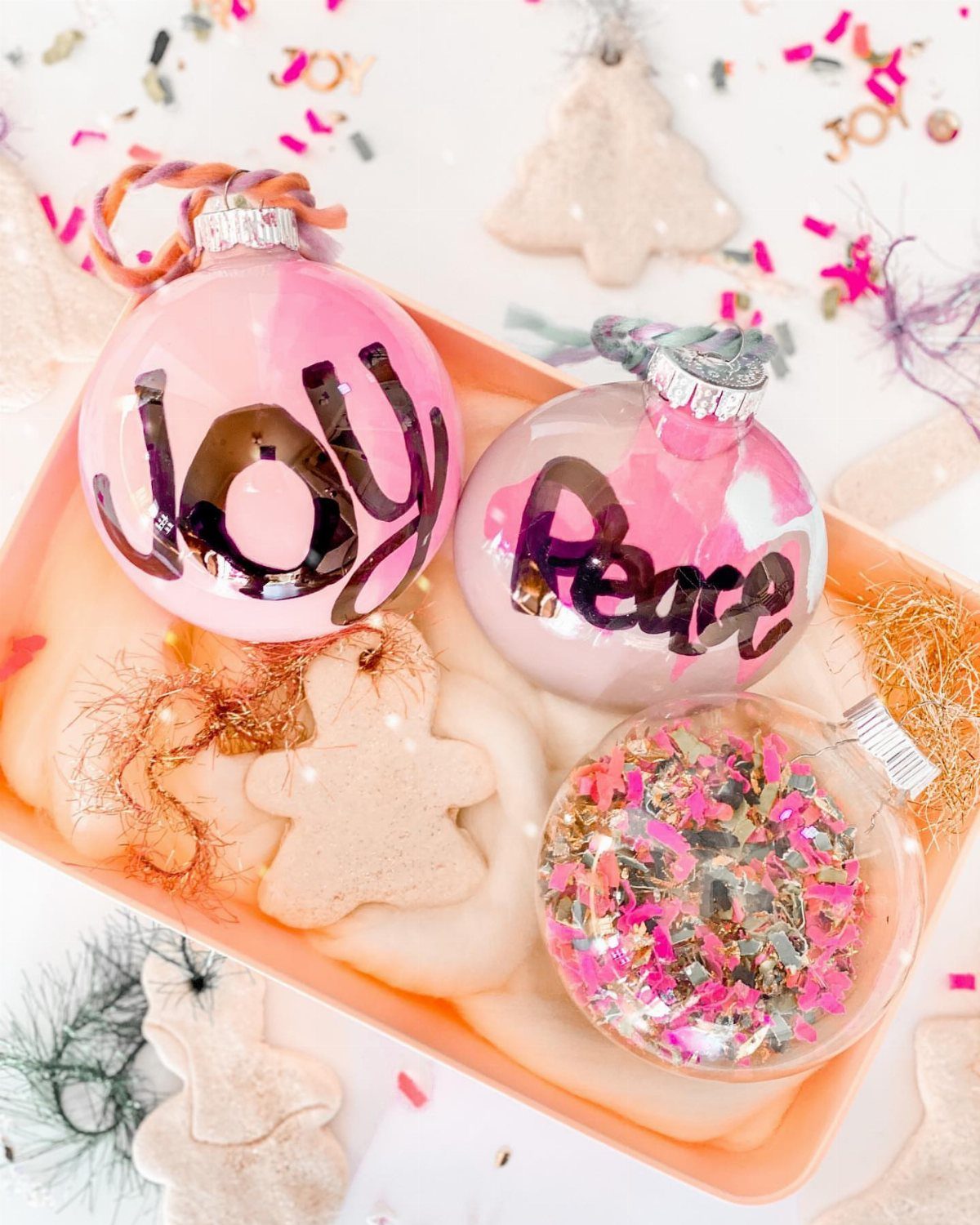 7 Ideas for Decorating Clear Ornaments with Glitter, Crayon & More 1 Three decorative glass ornaments resting in a tray, including one filled with pink and gold glitter.