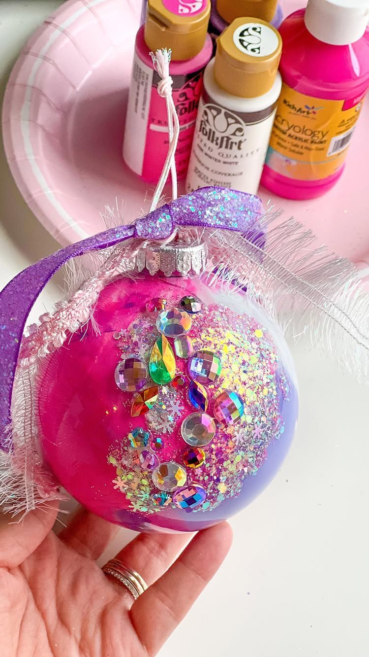 7 Ideas for Decorating Clear Ornaments with Glitter, Crayon & More 7 A hand holding a clear ornament decorated with pink paint, glitter, and colorful rhinestones.