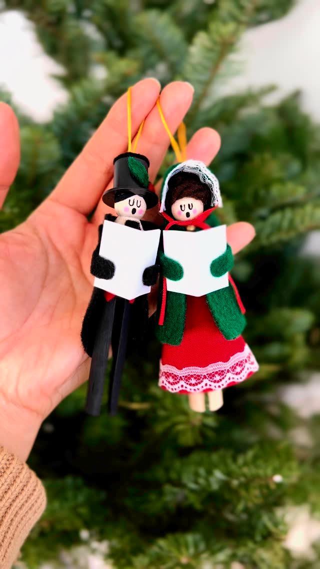 Two handmade clothespin ornaments shaped like people held in front of a green Christmas tree.