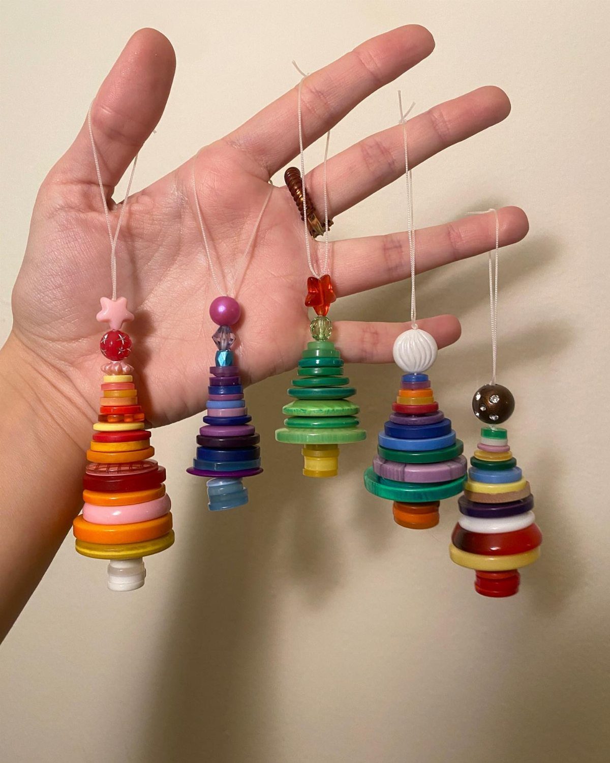 Five colorful button ornaments shaped like trees hanging from strings held in a hand.