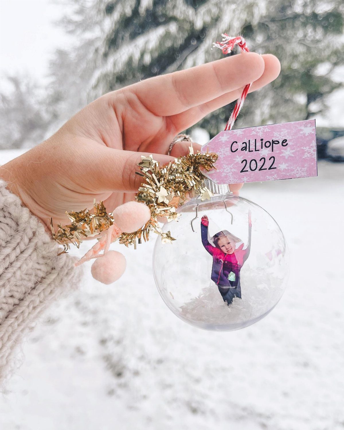A hand holding a clear snow globe ornament featuring a child's photo inside a snowy setting.