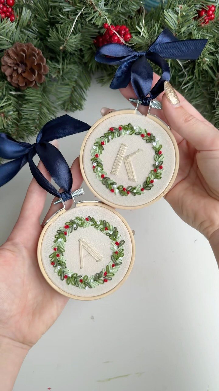 Two hands holding embroidered hoop ornaments with green wreaths and letters K and A.