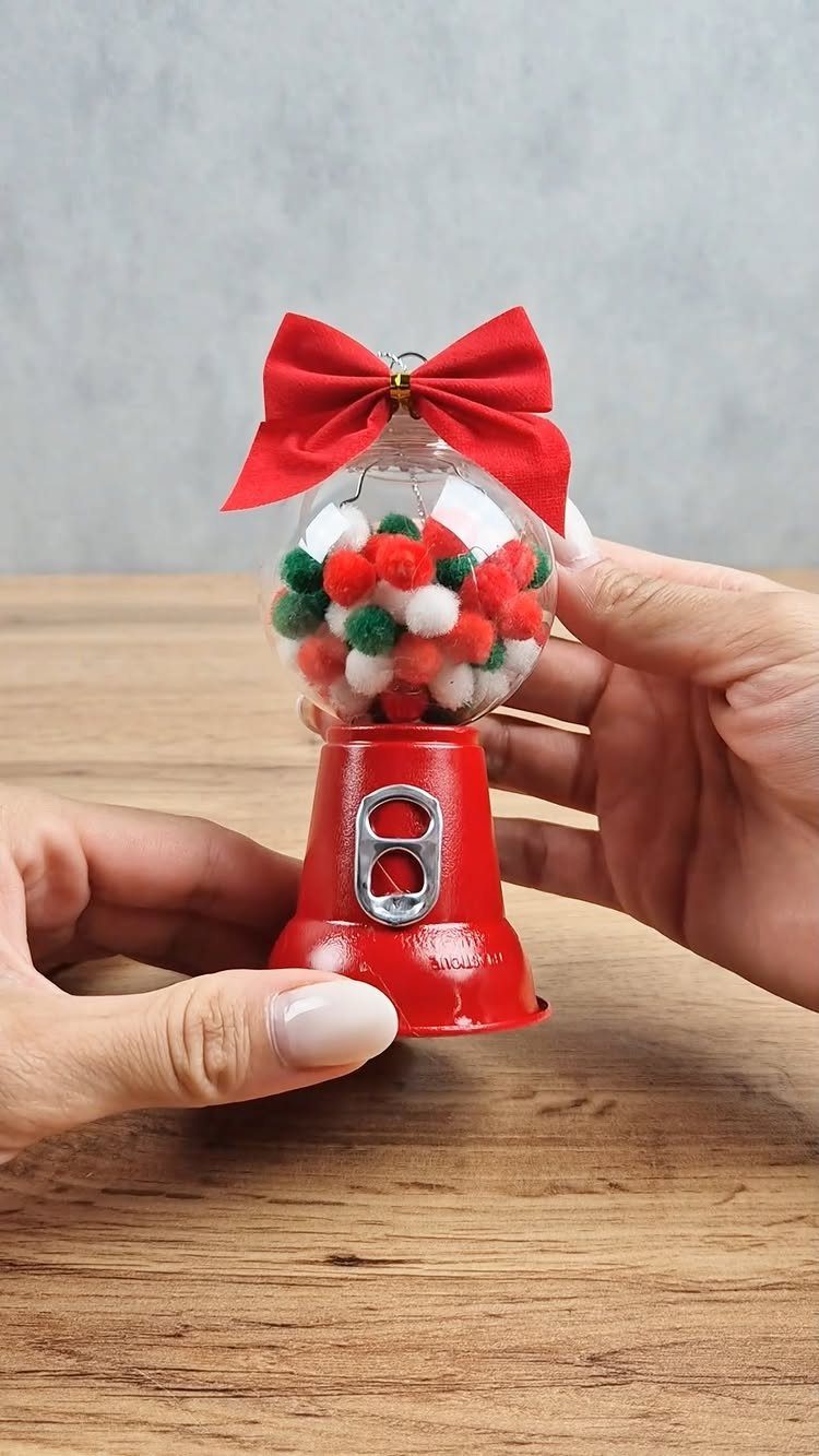 Hands holding a small red gumball machine ornament filled with colorful pom poms and a red bow.