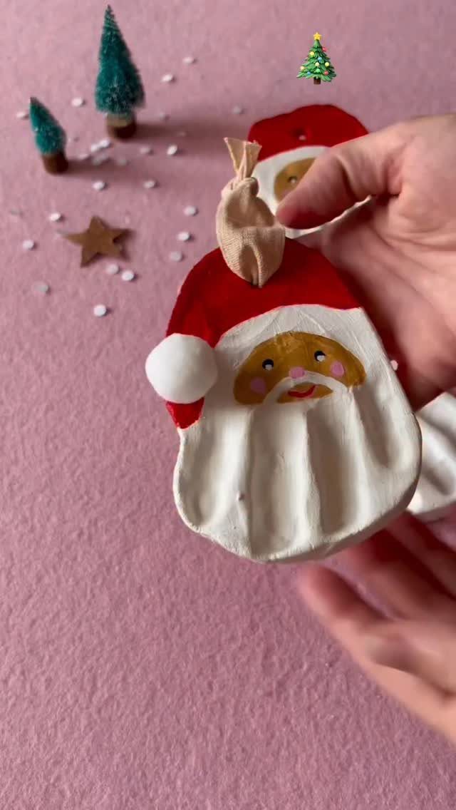 A hand holding a handmade Santa Claus ornament against a pink background with tiny trees.