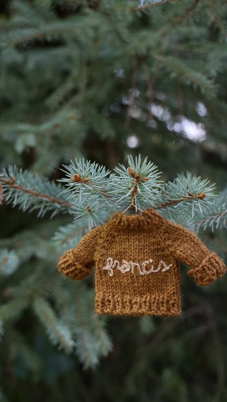 A tiny brown knitted sweater ornament with white embroidery hanging on a pine tree branch.