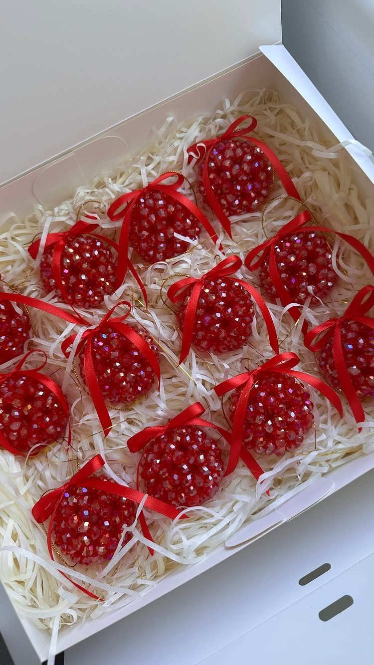 Red rhinestone-covered ornaments with satin bows resting in a white box with shredded paper.
