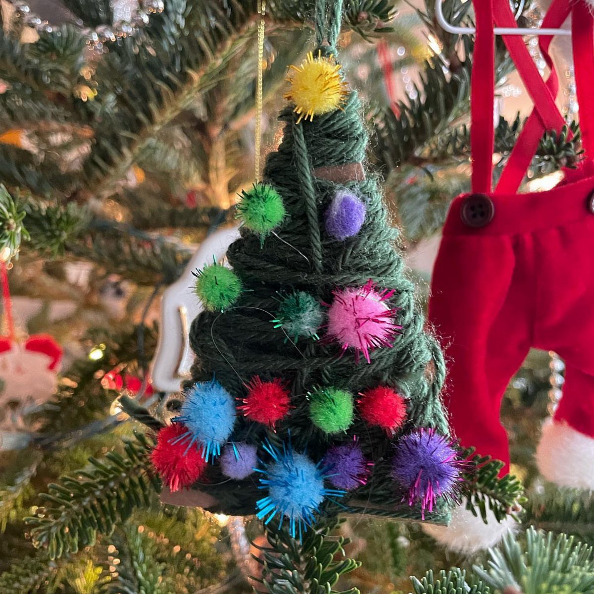 A green yarn tree ornament decorated with colorful fuzzy pom-poms hanging on a Christmas tree.
