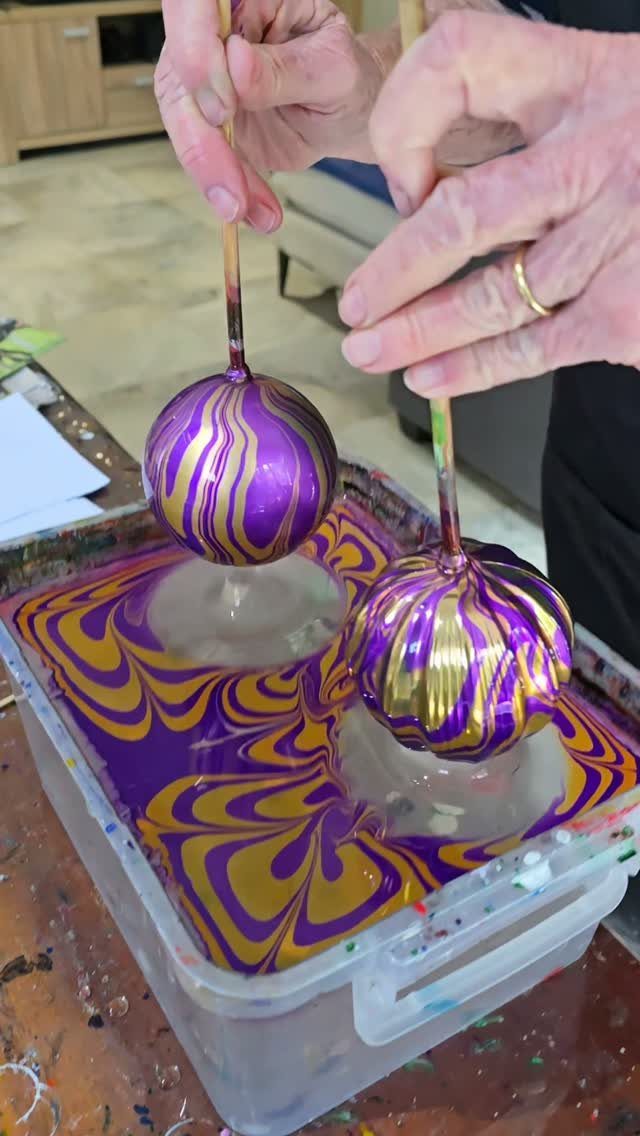 Hands holding two purple and gold swirled ornaments over a bin of colorful liquid.