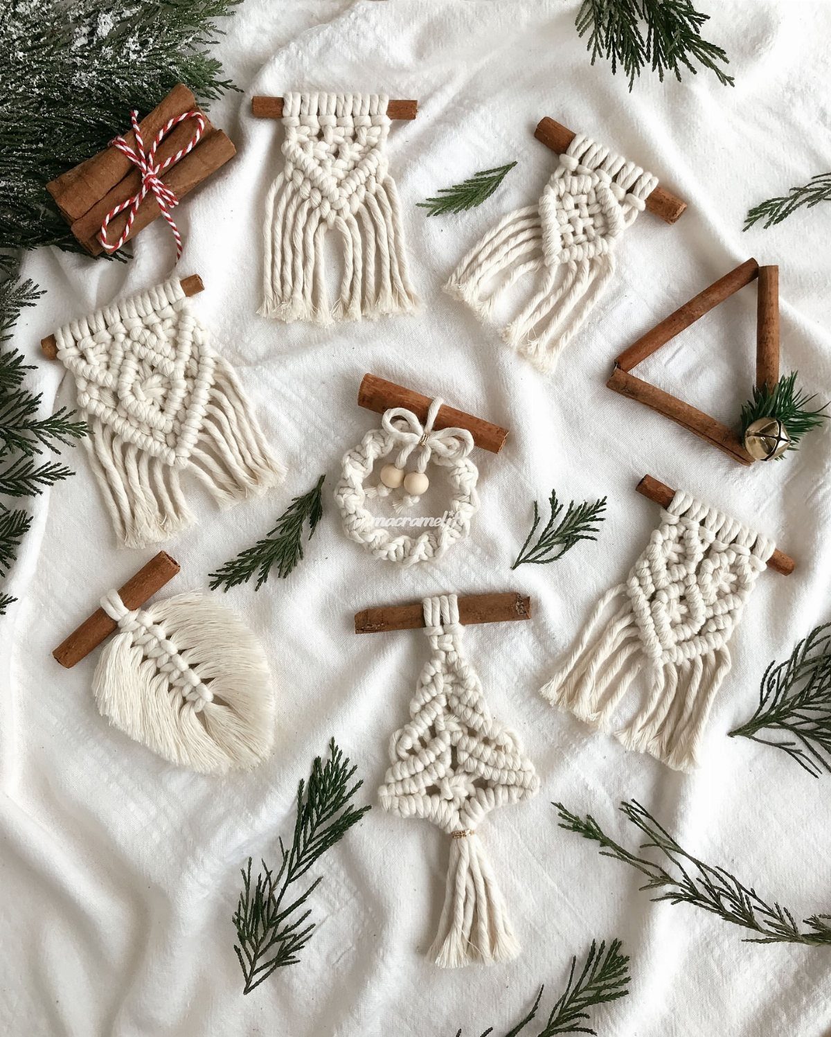 Macrame ornaments attached to cinnamon sticks arranged on white fabric with pine sprigs.