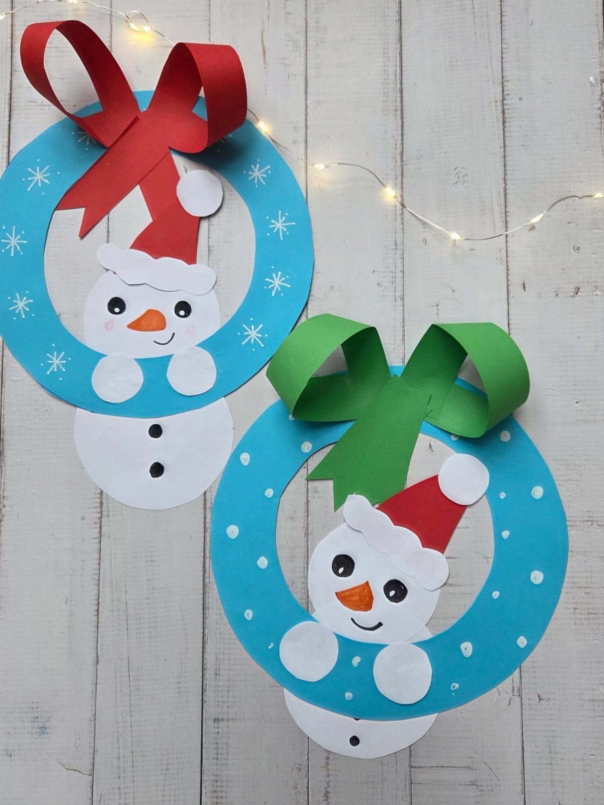 Two blue paper wreaths with snowman cutouts and large red and green bows on wood.