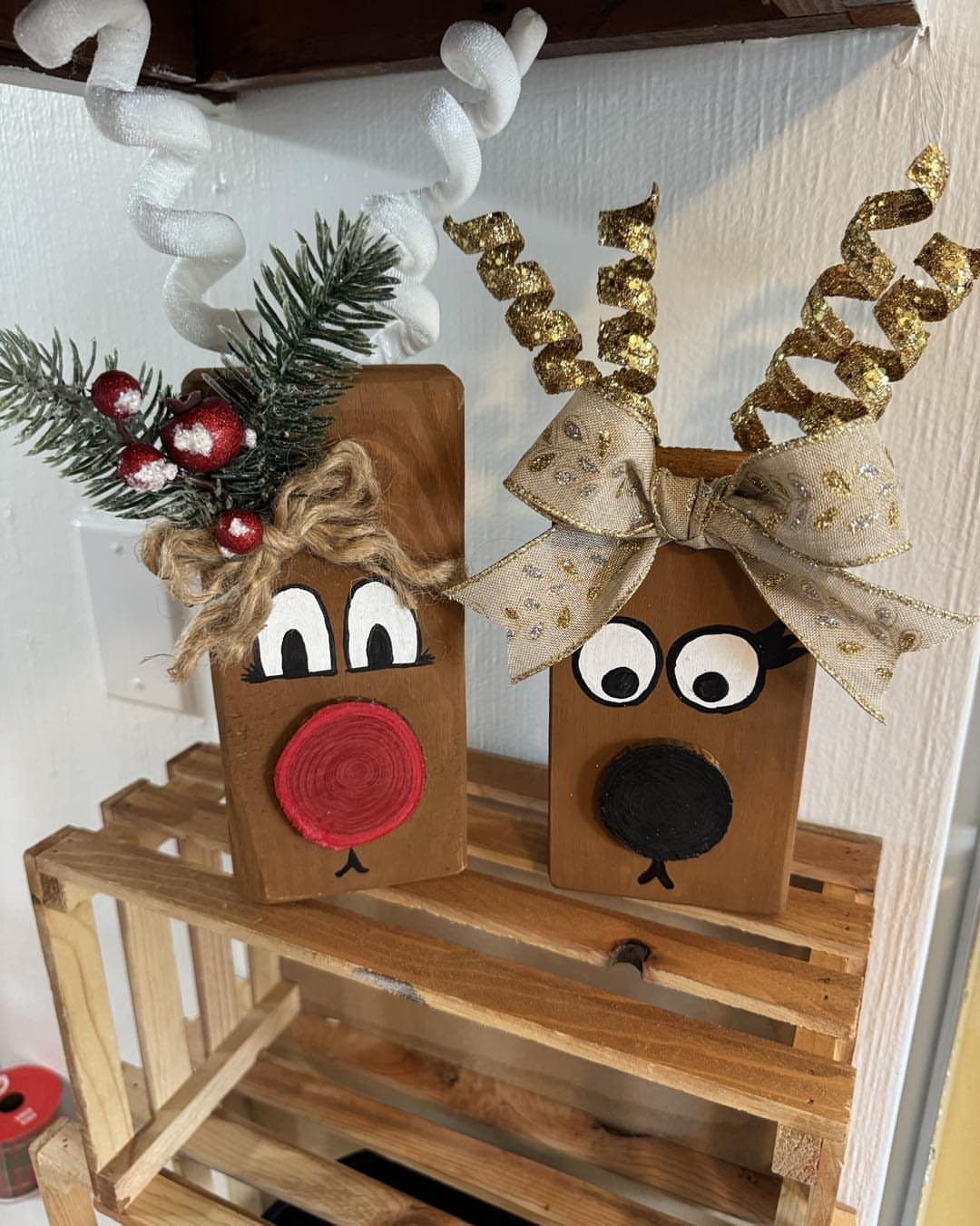 Two handmade wooden reindeer ornaments sitting on a small wooden crate against a white wall.