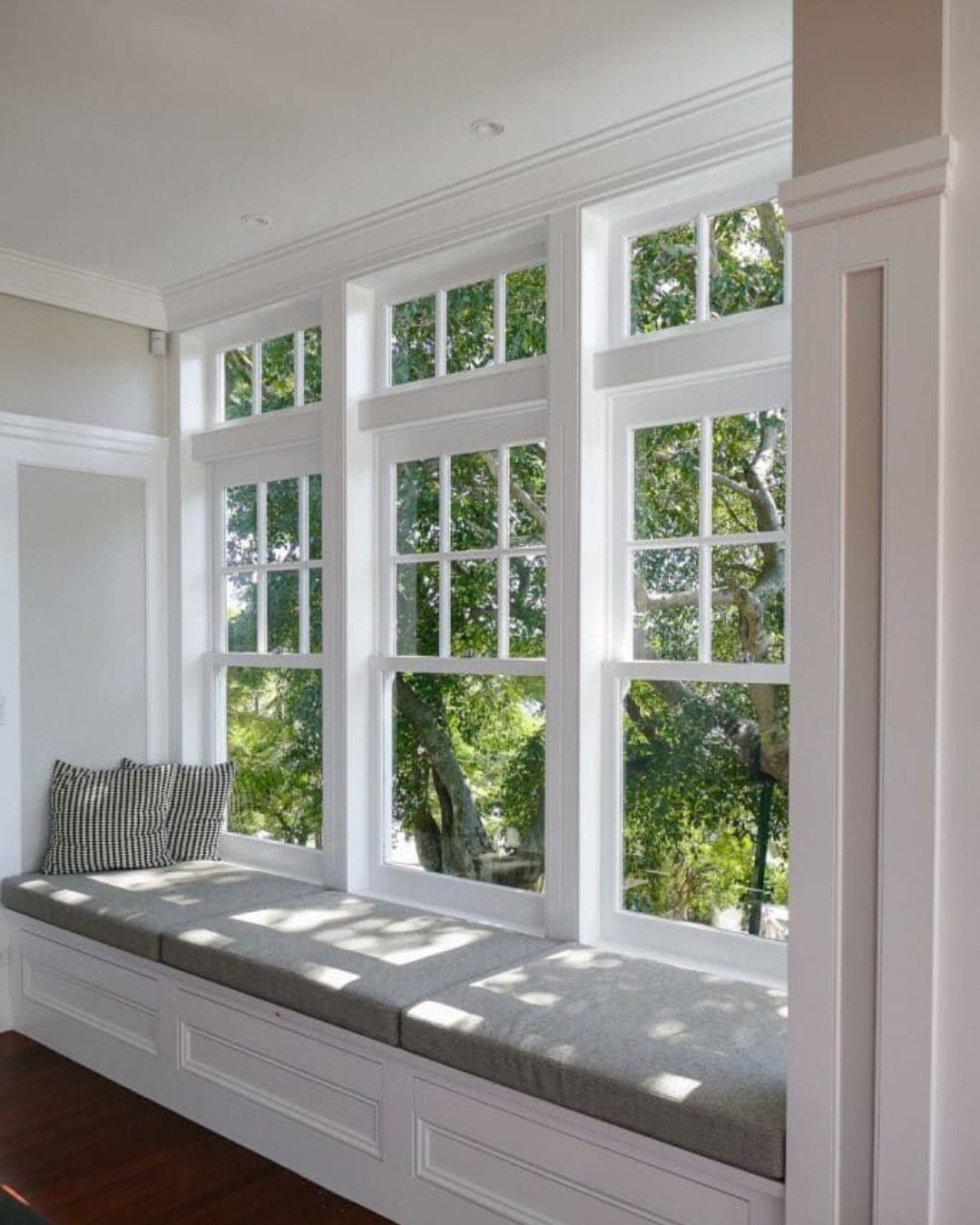 Front Window Design Ideas: 11 Looks Worth Stealing for 2026 3 Three large white double-hung windows with a built-in cushioned window seat overlooking green trees.