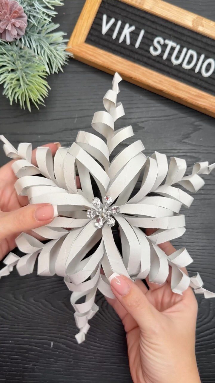 Homemade Christmas Decorations: 17 Easy Projects for 2026 9 Hands holding a white paper snowflake ornament with a rhinestone center on a dark table.