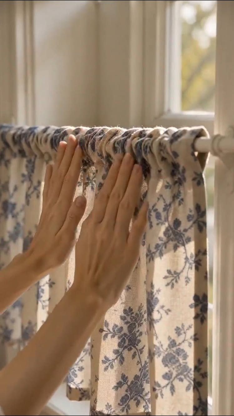 13 House Curtains Ideas for Every Style 6 Hands adjusting a short cream and blue floral patterned cafe curtain on a window rod.