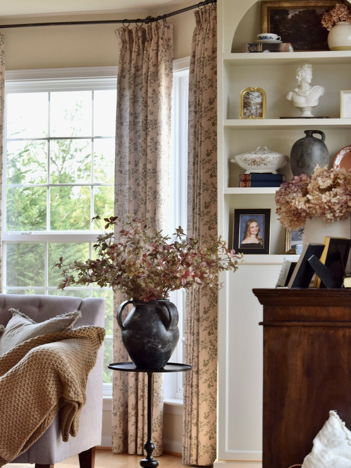 13 House Curtains Ideas for Every Style 9 Floral patterned curtains hanging by a large window next to a cozy armchair and bookshelf.