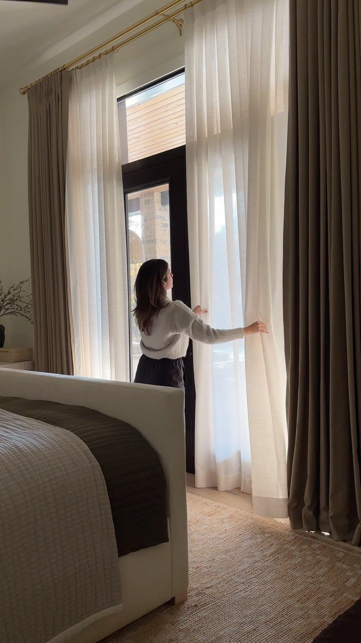 13 House Curtains Ideas for Every Style 1 A woman reaches toward layered white sheer curtains and heavy taupe drapes in a bedroom.